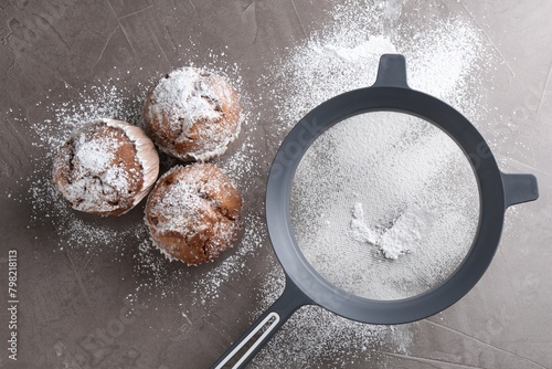 Sieve with sugar powder and muffins on grey textured table, flat lay photo