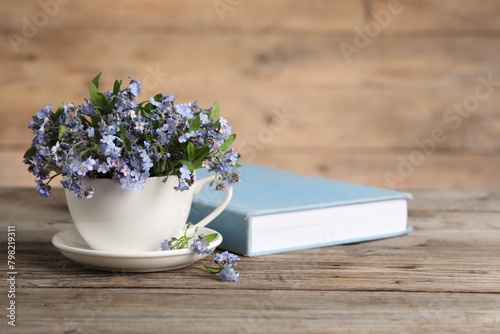 Beautiful forget-me-not flowers in cup  saucer and book on wooden table. Space for text