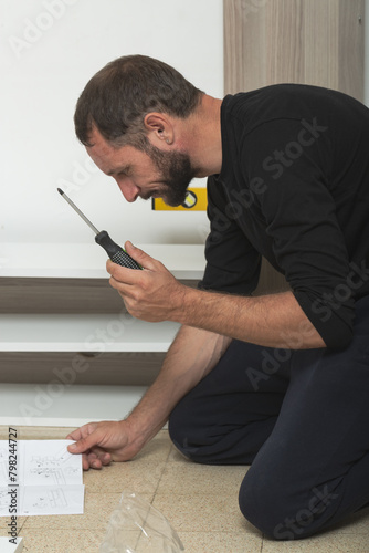 Man on the floor assembling furniture at home, holding a screwdriver and looking at assembly instructions. There is a partially assembled wooden shelves, a yellow level tool nearby. Screwdriver work photo