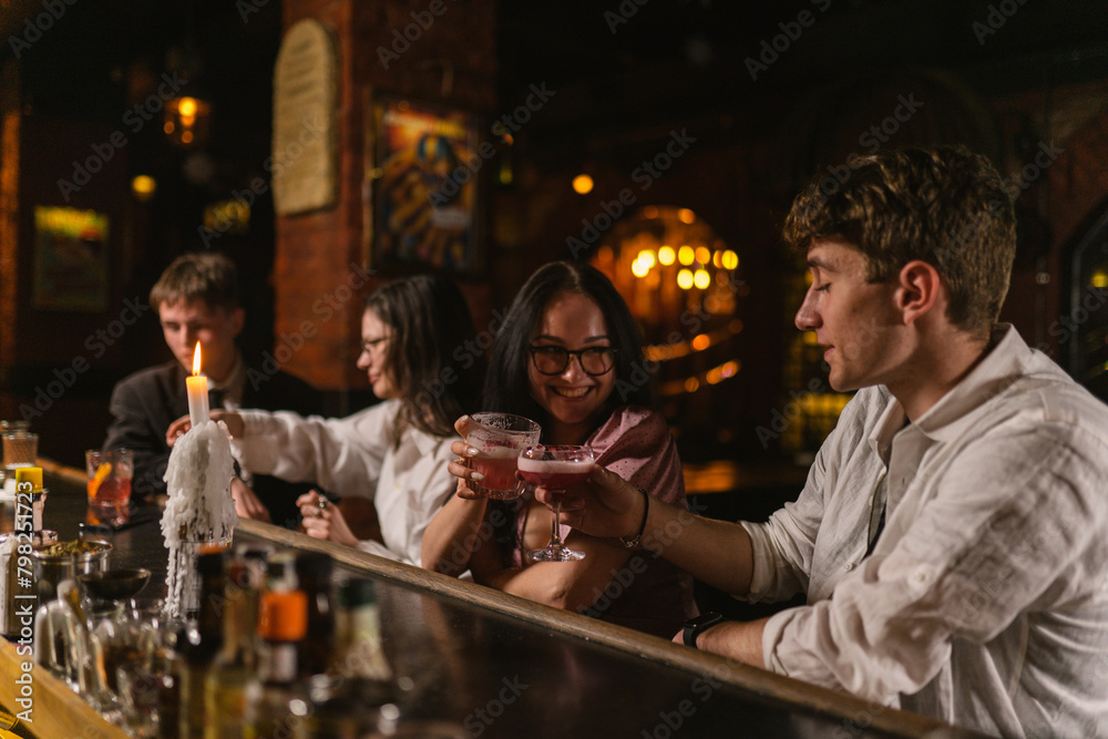 Guys and girls laugh and enjoying party together at bar. Young couple on date sit with candle at bar and raise cocktails in glasses.