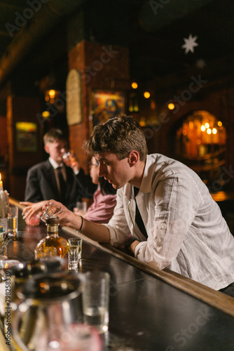 Depressed man lost job and drinks hard alcohol in bar with subdued light. Guy in white shirt sits among strangers at bar in cozy pub