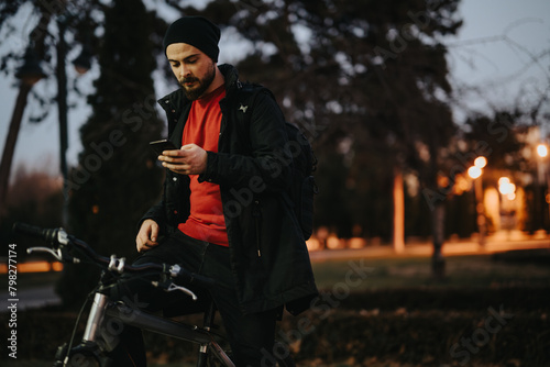 A young man stands by his bicycle in a park, using his smart phone as dusk settles in, with street lights glowing in the background.