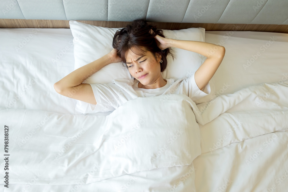 Thai woman wearing a white t-shirt can't sleep, stressed, scratching her head in discomfort on a white bed in a room with light shining through the window. Top view. Thai woman wearing a white t-shirt can't sleep, stressed, scratching her head in discomfort on a white bed in a room with light shining through the window. Top view.