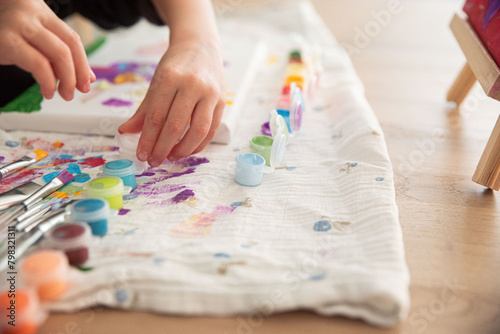 Cute little child painting with paintbrush and colorful paints. Kid hands start painting at the table with art supplies, top view