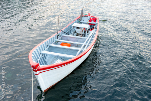 A white wooden traditional dory or small fishing vessel with red trim. The fishing boat has wooden oars, fishing rods, and a vintage outboard motor. The small craft vessel is moored in shallow water.