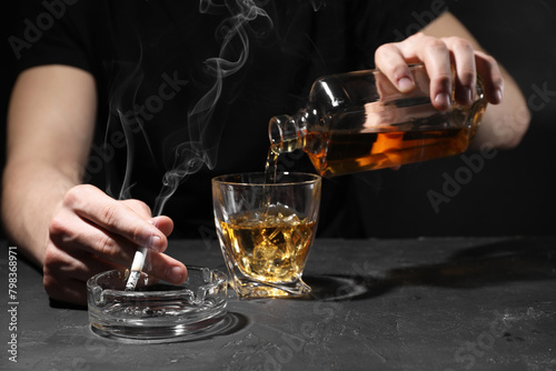 Alcohol addiction. Man with smoldering cigarette pouring whiskey into glass at dark textured table, closeup