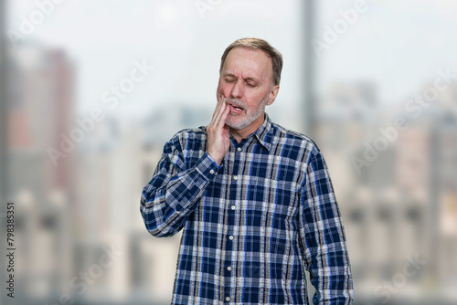 Mature office worker in checkered shirt having toothache. Blurred windows background. © DenisProduction.com