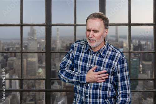 Portrait of a mature man having heartache touching his chest. Aged man having heart atack indoors. Checkered windows background with cityscape view. © DenisProduction.com