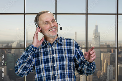 Portrait of smiling mature male call center customer support worker. Checkered window backgroud with cityscape view.