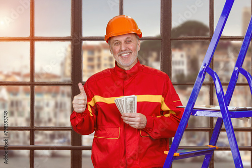 Portrait of smiling elder man with dollar baknotes showing like gesture. Handyman construction worker with his salary standing indoors.