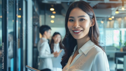 Smiling confident business leader looking at camera holding a digital tablet and standing in office at team meeting. Portrait of pretty Asian businesswoman with colleagues. AI Generated