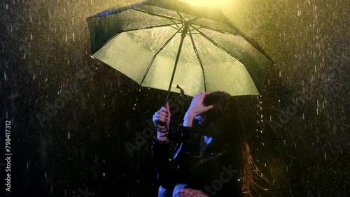 Woman runs through wet hair with hand standing under umbrella. Light of spotlight romanticizes scenery with lady in wet clothes in night rain photo