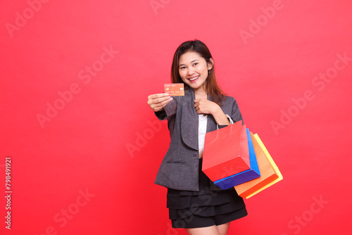 Asian office woman smiles carrying a shopping paper bag while holding a credit debit card to the front wearing a formal jacket with a red background. for transaction, lifestyle and business concepts