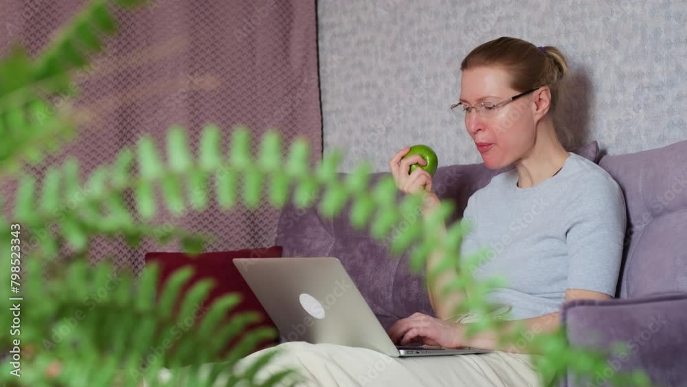 Happy caucasian woman using a laptop and eating green apple while sitting on couch in the living room. Remote employee, freelancer, programmer working at home.