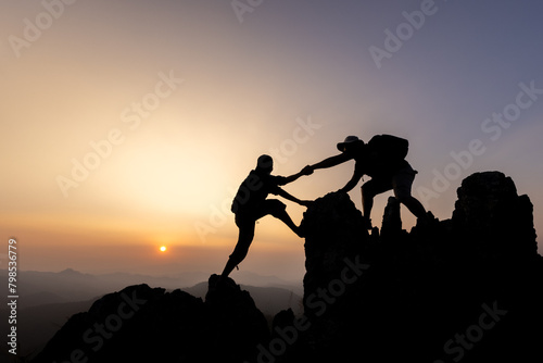 Silhouette of person hikers climbing up mountain cliff and one of them giving helping hand. People helping and, team work concept.