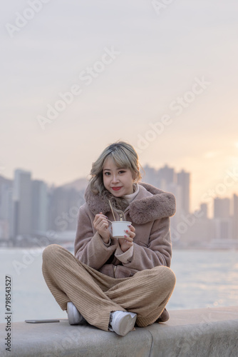 A young woman in winter clothes in her 20s who eats local food fish balls in the evening in a park with a view of buildings and the sea on Kowloon Island, Hong Kong
香港九龍島のビル群と海が見える公園で夕方にローカルフードのフィッシュボ