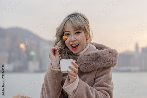 A young woman in winter clothes in her 20s who eats local food fish balls in the evening in a park with a view of buildings and the sea on Kowloon Island, Hong Kong
香港九龍島のビル群と海が見える公園で夕方にローカルフードのフィッシュボ