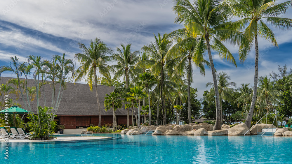 Picturesque boulders and coconut palms grow on the shore of the aquamarine swimming pool. Sun loungers are placed under sun umbrellas. A building with a tiled roof in the distance. Clouds, blue sky.