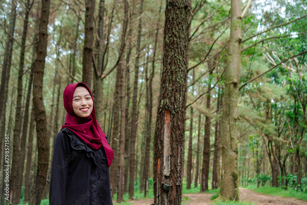 Javanese woman wearing a headscarf is standing in the middle of the forest facing the camera, against a background of trees with empty photocopy space