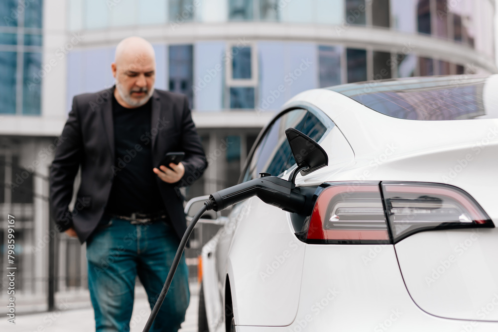 A businessman turns off an electric car with a full charge level at a charging station using an application on a smartphone. Concept of electric vehicles and modern technologies.