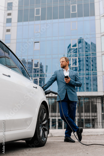 A business man uses a mobile phone while charging the battery of his electric car. A confident man in a business suit stands near a luxury car with a modern smartphone in his hands.  © Vladislav