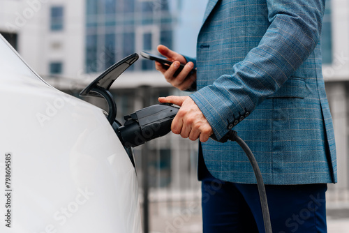 A middle-aged businessman charges an electric car from a charging station powered by renewable energy sources against the backdrop of the business center.
