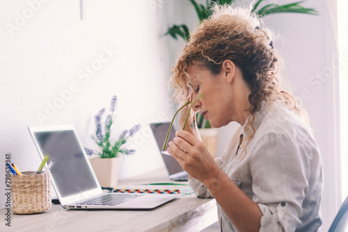Tired stressed middle age business woman suffering from neckpain working from home office sitting at table. Overworked adult lady massaging neck feeling hurt pain from incorrect posture. Lifestyle photo