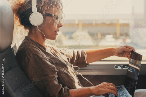 Mid age woman using computer inside transport bus vehicle during business or vacation travel. Modern work digital lifestyle. Female people with headphones enjoy transport. Traveler lady with laptop photo
