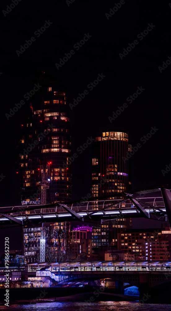 Obraz premium Millennium Bridge illuminated at night, with tourists walking and a night train stopped on Blackfriars Bridge with skyscrapers behind with illuminated windows and the London Eye in the background.