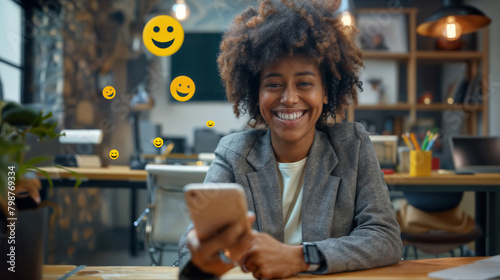 A young smiling black woman holding a mobile phone in the office with emoji smiles flying around her. The person receives a pleasant message
