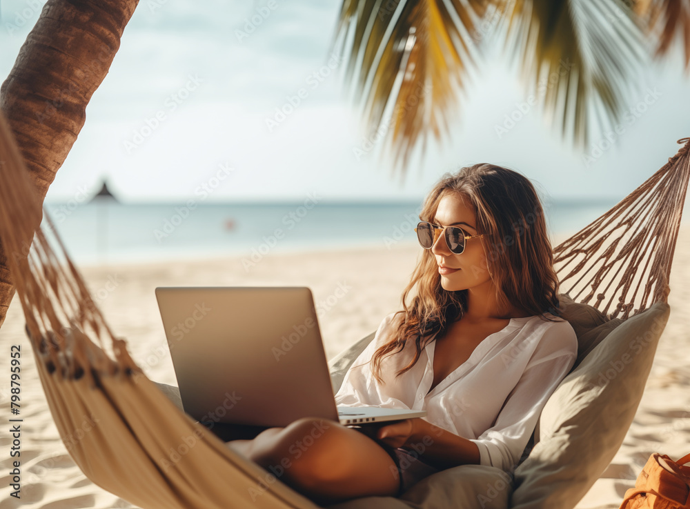 Enjoying work during summer vacation. Young happy woman, successful female freelancer in straw hat using laptop and smilingwhile relaxing in the hammock on the tropical beach. Distance job