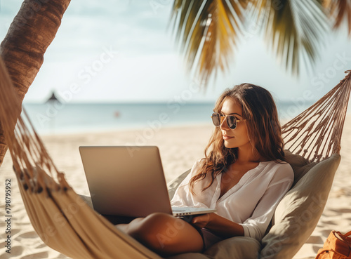 Enjoying work during summer vacation. Young happy woman, successful female freelancer in straw hat using laptop and smilingwhile relaxing in the hammock on the tropical beach. Distance job photo
