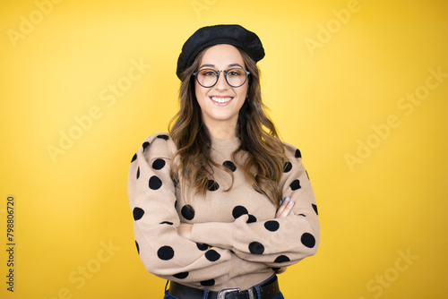 Young beautiful brunette woman wearing french beret and glasses over yellow background with a happy face standing and smiling with a confident smile showing teeth with arms crossed