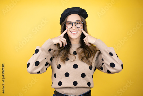 Young beautiful brunette woman wearing french beret and glasses over yellow background very happy and excited making winner gesture with raised arms, smiling and screaming for success.