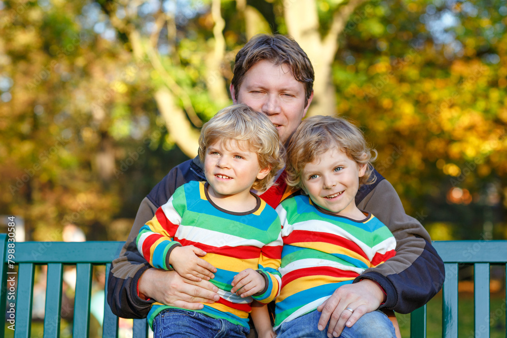 Fototapeta premium Two little kid boys and young father sitting together in colorful clothing on bench. Cute healthy children, siblings and their dad having fun in autumn park on warm sunny day. Happy family of three.