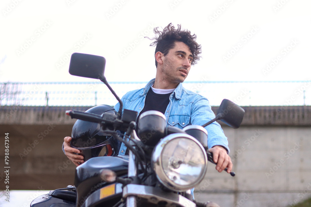 Portrait of curly haired Caucasian young man holding his motorcycle and helmet, looking to the side with bridge in the background. Lifestyle concept