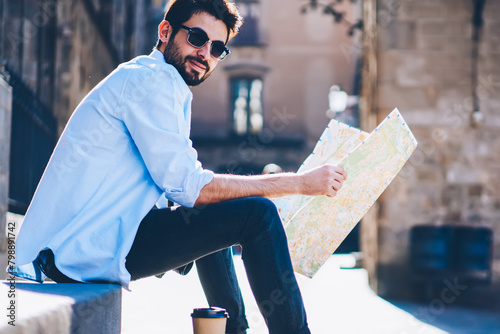 Portrait of cheerful bearded tourist in sunglasses sitting outdiirs on street of architectural city with old constructions while holding travel map for searching destination of showplaces photo