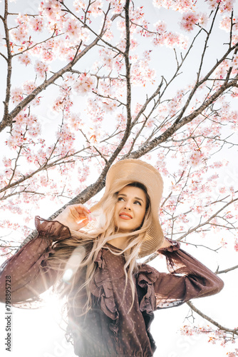 Young pretty woman in hat standing in blooming sakura tree