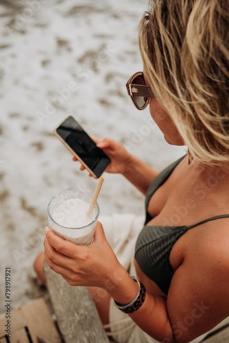 Young woman on the beach using mobile phone and drinking cocktail