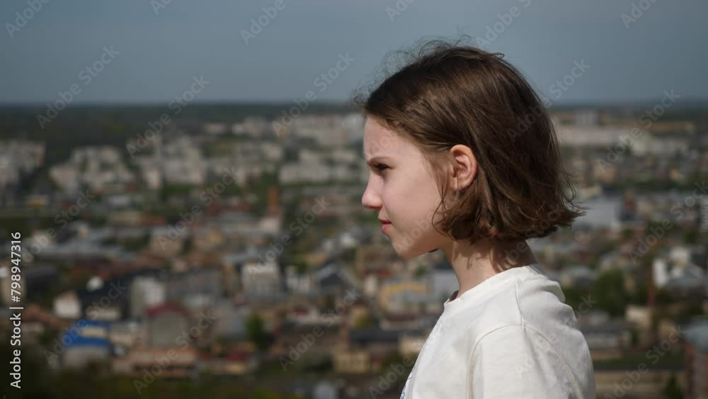 Portrait of pensive teenage girl standing high on mountain against backdrop of city. Child girl looks into distance, wind blows her hair. Feeling lightness and freedom during hike in mountains.