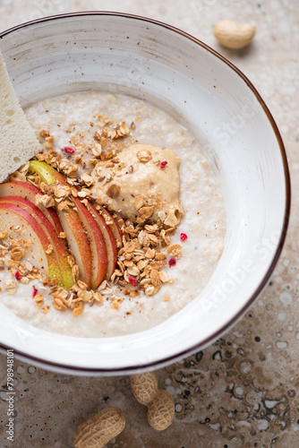 Bowl of oatmeal porridge with peanut butter and fresh apple, vertical shot on a beige granite background, middle close-up