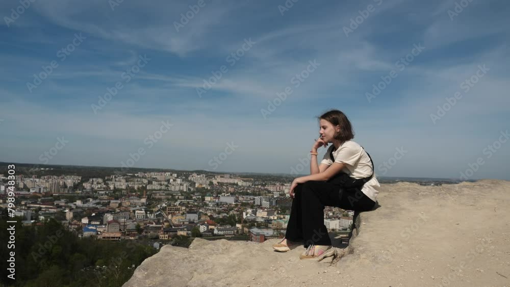 pensive teenage girl sitting high on mountain against backdrop of city. Child girl looks into distance, wind blows her hair. Feeling lightness and freedom during hike in mountains.