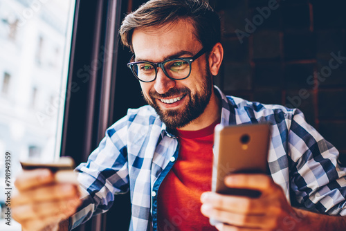 Cheerful bearded student with banking card in hands making payment on website on smartphone via internet connection.Positive young man looking at business card holding phone for money transaction photo