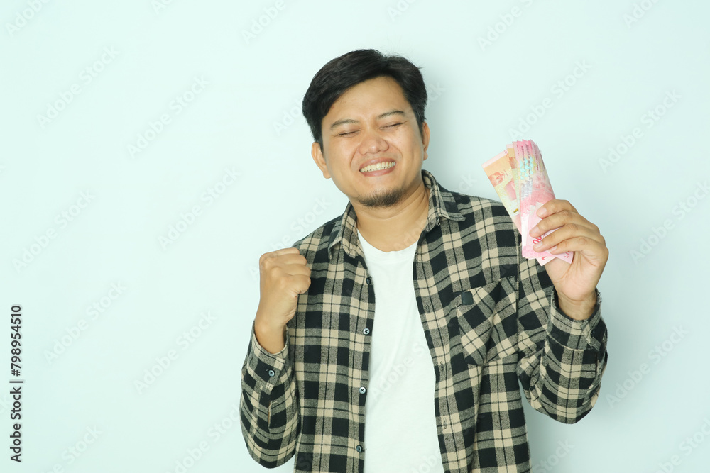 Young Asian man showing excited face expression with fist clenched and close the eyes while holding a wallet full of money Young Asian man showing excited face expression with fist clenched and close the eyes while holding a wallet full of money