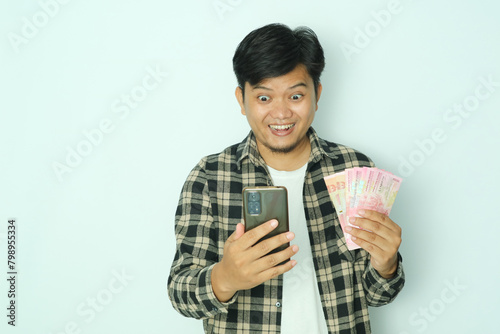 Young Asian man wearing brown flannel shirt holding money showing excited expression when looking to his mobile phone photo