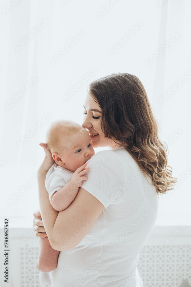 A happy young mother and a newborn baby smile, hug and kiss on a white isolated window background, a loving family at home A happy young mother and a newborn baby smile, hug and kiss on a white isolated window background, a loving family at home