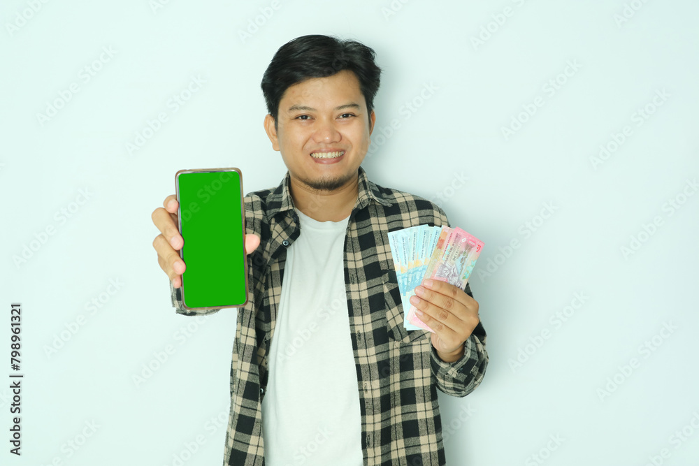 Young Asian man wearing brown flannel shirt smiling happy while showing blank mobile phone screen and holding paper money Young Asian man wearing brown flannel shirt smiling happy while showing blank mobile phone screen and holding paper money