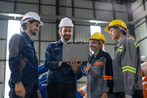 A diverse group of smiling industrial professionals collaborates over a laptop on the factory floor, embodying teamwork and technological integration in manufacturing.
