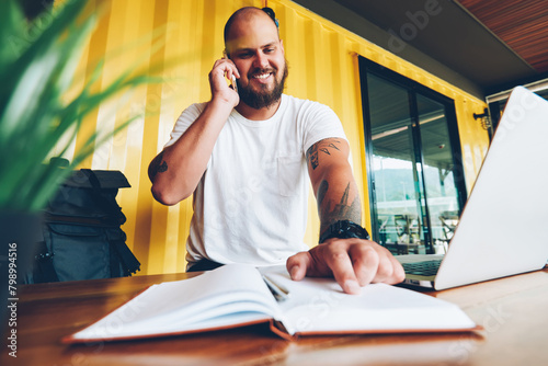 Cheerful hipster guy pointing on notes in notebook share information during mobile phone talk. happy male student calling to friend consulting about homework sitting in college campus with laptop © BullRun