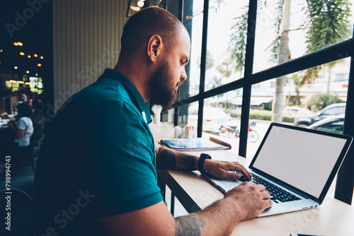 Young bearded guy sitting in cafeteria with laptop computer typing text for publication in blog,skilled male freelancer doing distance job via netbook with mock up screen and wireless internet photo